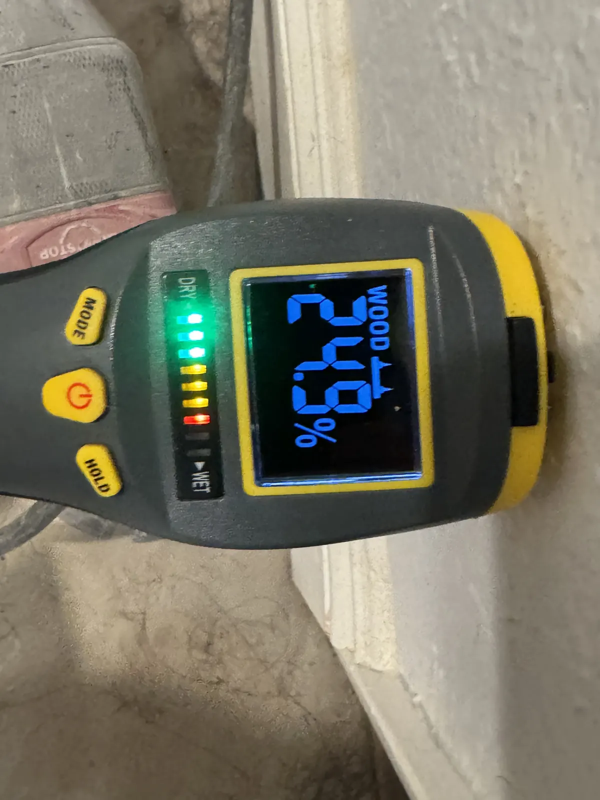Moisture meter held against the base of a wall, reading 64.2