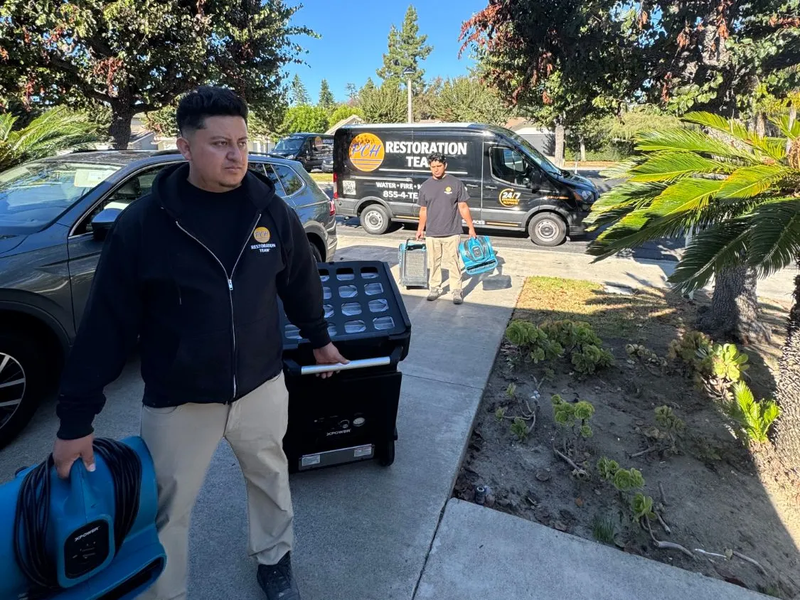 PCH Restoration Team technicians carrying an air mover and HEPA air scrubber up a homeowner's driveway, response van parked behind them