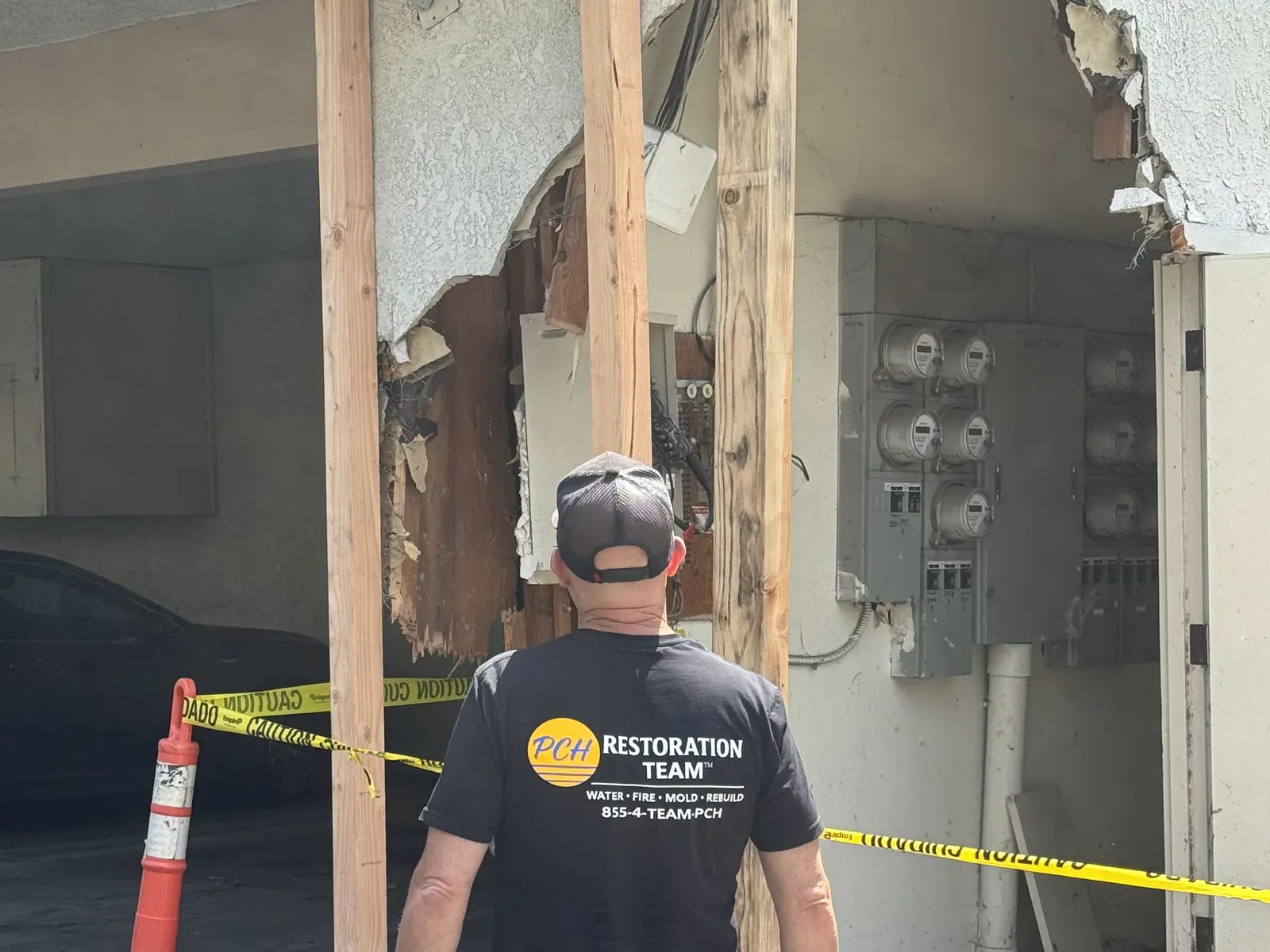 PCH Restoration Team technician inspecting a damaged multi-unit carport wall behind caution tape and safety cones